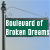 Green street sign with white text reading 'Boulevard of Broken Dreams' on a light blue background, mounted on a dark pole.
