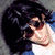 Pixelated close-up of a person with dark hair wearing sunglasses and a white shirt against a blue background.
