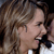 A pixelated close-up of a person with long brown hair holding a microphone near their mouth, wearing a white garment with dark straps.