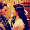 A static image shows two people in period attire standing close together in a warmly lit interior with ornate architectural details.