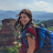 Pixelated static image of a person with long brown hair wearing a red and blue garment, standing outdoors with a blurred background of blue sky and greenery.
