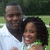 Two people stand close together outdoors with greenery in the background; one wears a light-colored shirt and the other a green top.