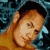 Pixelated static image of a person with light brown hair and blue eyes against a dark teal background, rendered in low-resolution 8-bit style with visible dithering and color banding.