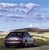 Pixelated image of a dark-colored sedan on a road under a blue sky with scattered clouds, viewed from a low angle.