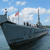 A pixelated ship with dark hull and white smoke against blue sky, featuring a red flag on a mast and multiple smokestacks.