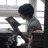 A person with short dark hair reads a book in a room with a window, viewed from a side angle.
