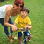 A child in a yellow shirt and blue pants rides a bicycle with an adult behind, both on grass.
