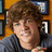Pixelated image of a person with brown curly hair wearing dark clothing against an orange background with framed items.