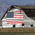 A pixelated image of a white barn with a black roof, featuring a large American flag on its side, set against a blue sky and brown ground.