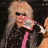Pixelated image of a person with light-colored hair wearing dark sunglasses and a black jacket over a pink tie, holding an object near their face.