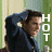 A pixelated image of a person with dark hair wearing a dark blue jacket, standing against a striped background with green and beige vertical stripes, with the word "HOT" in white text vertically centered.