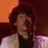 A person with curly brown hair wearing a white jacket over dark clothing, holding a microphone against an orange and red background.