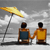 Two figures sit side-by-side holding hands under a yellow beach umbrella on a gray background.