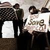 A static image shows a person holding a sign reading 'God Save America!' with an American flag draped over a structure in the background.