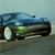 A pixelated blue car with green accents on a gray road under a light blue sky, viewed from a low angle.