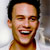 Pixelated close-up of a smiling person with dark hair and light skin, wearing a brown shirt against a blurred background.