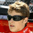 A pixelated image of a person with short light brown hair wearing dark sunglasses and a red garment, set against a red background with white text elements.
