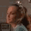 A person with light brown hair in a ponytail wears a light blue top and a small white headpiece, positioned against a brown wall with a framed picture.