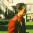 Pixelated static image of a person in an orange jacket and dark shirt standing outdoors with greenery and buildings in the background.