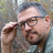 Pixelated static image of a person with short dark hair, glasses, and a beard, wearing a dark shirt, with a blurred background of muted colors.