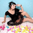 A pixelated image of a woman in a black dress sitting on the ground with flowers, her right hand raised to her head.