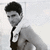 Pixelated grayscale image of a person with short dark hair wearing a light jacket over a white shirt and dark tie, standing against a plain background.