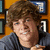 Pixelated image of a person with brown curly hair wearing a dark blue shirt, set against a background with framed items on an orange wall.