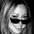 Pixelated grayscale portrait of a person with long hair wearing dark sunglasses and a slight smile, cropped to show head and shoulders against a dark background.