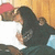 Two figures in a close embrace, one wearing a red cap and white shirt, the other with long dark hair and dark clothing, against a wooden background.