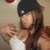 A person with long brown hair wearing a black cap and white top, holding a white object near their chest in a static pose.