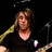 A person with shoulder-length brown hair wearing a black V-neck shirt plays an electric guitar against a dark background with microphones.