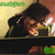 A person in dark clothing holds a green object near their face, with a blurred background and green text at the top.