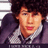 Pixelated static image of a person with dark hair wearing a dark jacket over a light collared shirt, red lips, and a neutral expression.
