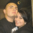 Two people in dark clothing embrace; one wears a cap with a yellow emblem, the other has short dark hair and a white collar.