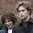 Two people with brown hair stand side-by-side, one in a dark jacket over a white shirt, the other in a dark jacket over a light blue shirt, against a blurred background.