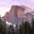 Pixelated landscape with a large brown rock formation under a light purple sky, dark green trees in the foreground, and a bright white light source on the left.