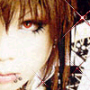A close-up of a person with brown hair and red eyes, wearing white clothing with black patterns, against a dark background.