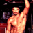 A pixelated image of a shirtless man with a muscular build, raised right arm, and neutral expression against a dark background with blurred red and purple elements.