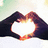 Two hands form a heart shape with fingers, creating a bright white heart-shaped void against a soft green and white background.