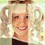 Pixelated image of a smiling person with light hair wearing a dark top against a light background with faint circular patterns.
