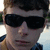 Pixelated close-up of a person wearing dark sunglasses and a dark cap, with light-colored hair visible at the sides, against a blurred outdoor background.