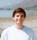 A person in a white long-sleeve shirt stands against a beach background with water and mountains, with animated text changing near the lower right.