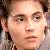 Pixelated close-up of a person's face with light skin tone, brown hair, and neutral expression against a blurred background.