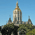 Pixelated image of a tall, light-colored tower with a spire, surrounded by smaller structures and greenery under a clear blue sky.