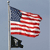 Static pixelated image of a waving American flag on a pole with a black triangular flag below it against a light blue background.