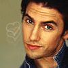 A close-up portrait of a person with dark hair wearing a blue collared shirt, set against a light brown background with faint heart outlines.
