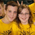 Two people in yellow shirts pose closely together, one making a peace sign with their right hand, against a dark background.