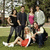 Group of people in outdoor setting with trees and grass, some standing, some crouching or sitting on ground.