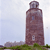 Pixelated image of a tall brown tower with a rounded top, set against a light blue sky with green ground at the base.