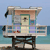 Pixelated static image of a beach house with a lifeguard tower structure, featuring a light brown roof, green trim, and blue and purple accents, set against a light blue sky and ocean background.